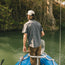A man in a pearl snap shirt and cap stands at the edge of a blue inflatable boat, fishing on a calm river surrounded by lush greenery and tree roots along the riverbank.