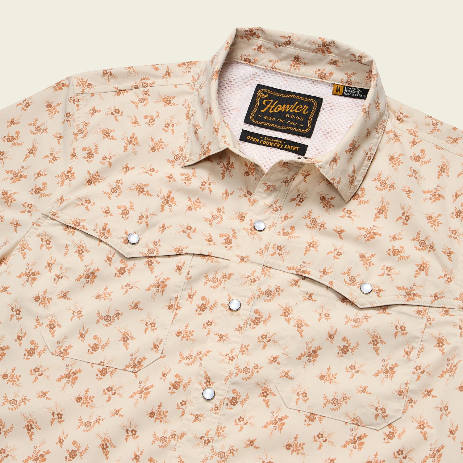 A beige, long-sleeve button-up shirt with a small orange floral print, crafted from nylon-poly fabric. Features pearl snaps and western-style chest pockets. The label reads Howler Bros and indicates a medium size.