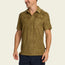 A man in a modern fit, olive green patterned vintage polo with a chest pocket, smiling and looking to the side. He pairs it with dark navy shorts.