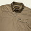 A khaki-colored tech polo with a button-up placket and buttoned chest pocket. Made from recycled polyester, it features a subtle textured pattern, Howler Bros. label inside the collar, and a small Howler tag on the pocket.