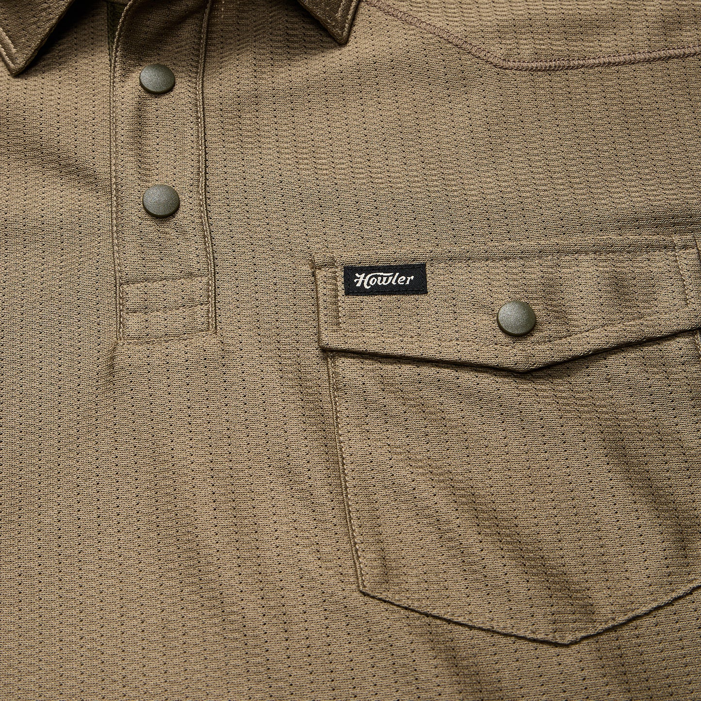 Close-up of a khaki textured shirt crafted from recycled polyester, featuring snap buttons, a flap chest pocket with a snap closure, and a small black Howler brand tag above the pocket.