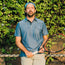 A man in a blue Tech Polo made from breathable recycled polyester stands outdoors in front of leafy greenery, holding a tennis racket and leaning on a chain-link fence.