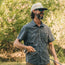 A man with a beard and sunglasses, wearing a patterned technical utility shirt with a vented rear yoke and a cap, stands outdoors holding a fishing rod, surrounded by green trees and sunlight.