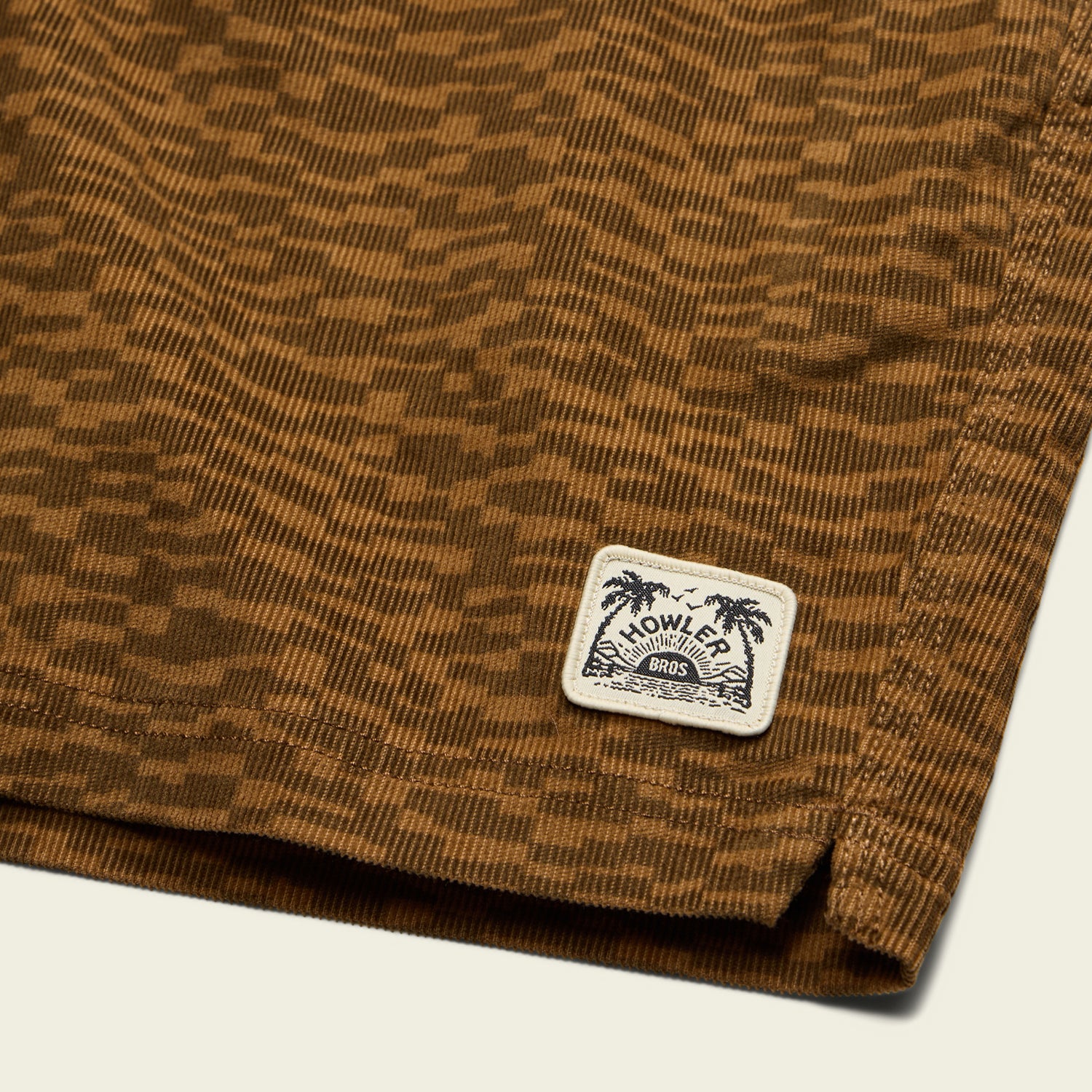 Close-up of brown stretch cotton corduroy with a geometric pattern, featuring a sewn white label with palm tree graphics and the text “Howler Bros”—a signature touch on their Pressure Drop Cord Shorts comfort shorts.