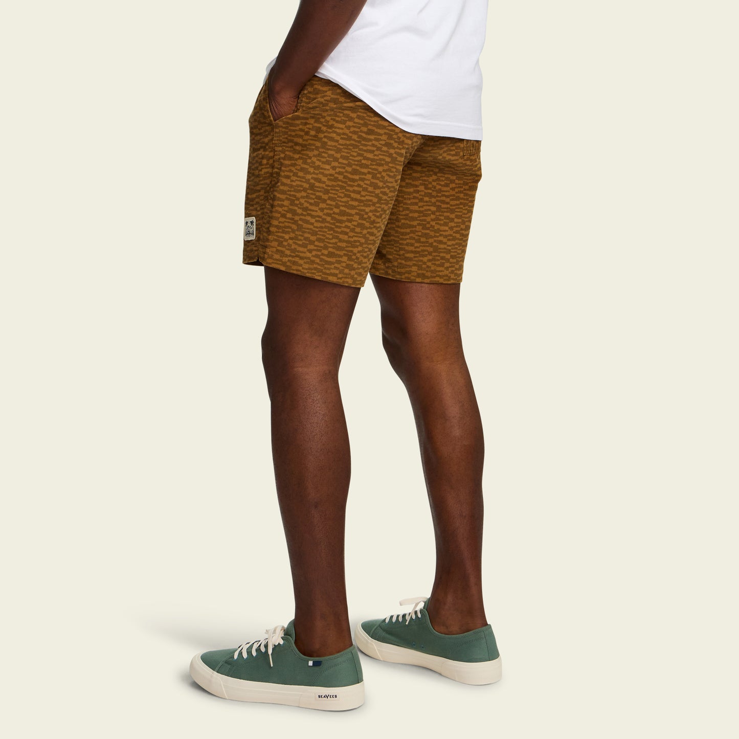 A person stands with hands in the pockets of mustard yellow Pressure Drop Cord Shorts, made from stretch cotton corduroy, wearing a white shirt and green sneakers with white soles, against a plain light background.