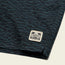 Close-up of dark blue and black patterned stretch cotton corduroy fabric with a small rectangular patch featuring palm trees and the words Howler Bros stitched near the edge, part of the Pressure Drop Cord Shorts collection.