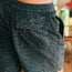 Close-up of a person wearing Pressure Drop Cord Shorts in patterned dark green and black stretch cotton corduroy with an elastic waistband, featuring a back pocket with a button. The background is blurred, with a green can visible.