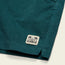 Close-up of dark teal stretch cotton corduroy, likely comfortable shorts such as the Pressure Drop Cord Shorts, with a rectangular cream Howler Bros logo patch and palm tree illustration near the hem.