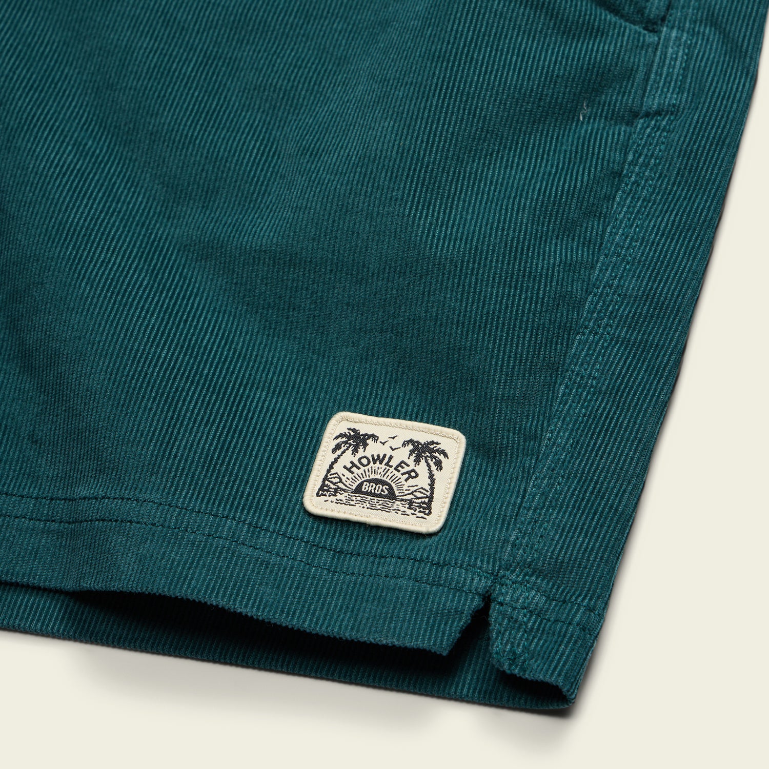 Close-up of dark teal stretch cotton corduroy, likely comfortable shorts such as the Pressure Drop Cord Shorts, with a rectangular cream Howler Bros logo patch and palm tree illustration near the hem.