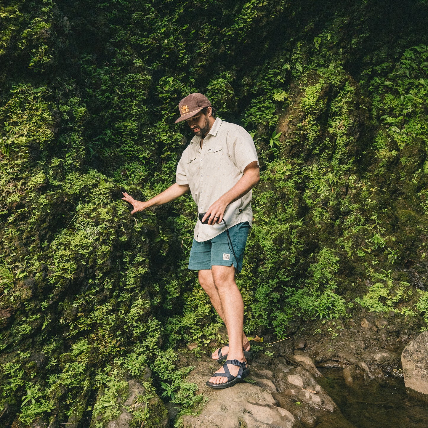 A man wearing a cap, beige shirt, and Pressure Drop Cord Shorts made from stretch cotton corduroy walks carefully on rocky ground next to a moss-covered wall in a lush outdoor setting.