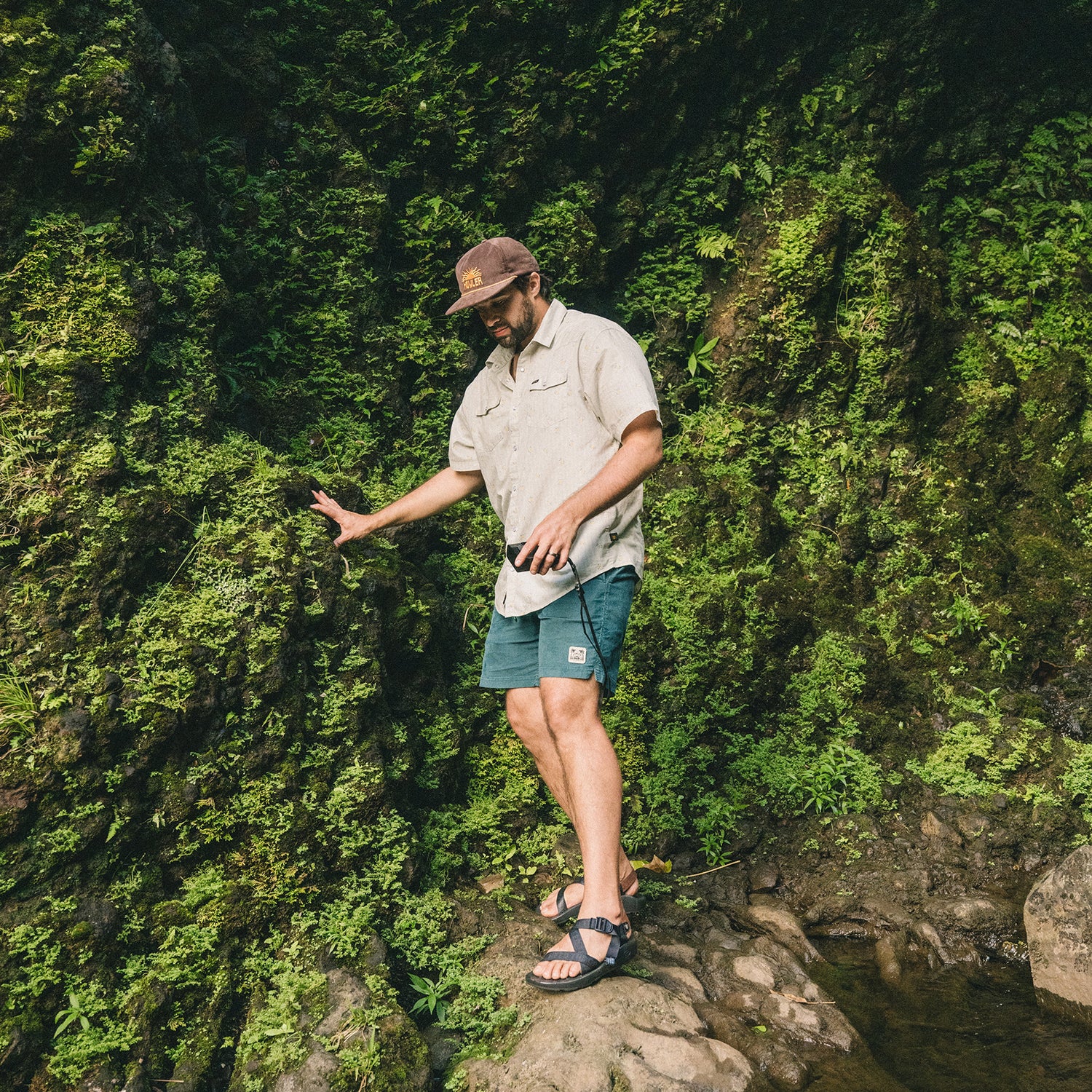 A man wearing a cap, beige shirt, and Pressure Drop Cord Shorts made from stretch cotton corduroy walks carefully on rocky ground next to a moss-covered wall in a lush outdoor setting.