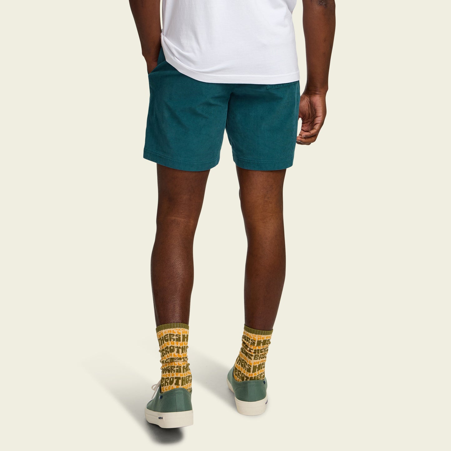 A person stands facing away, wearing a white T-shirt and comfortable shorts—teal Pressure Drop Cord Shorts made from stretch cotton corduroy—paired with yellow patterned socks and green sneakers against a plain light background.