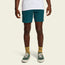 A person stands against a light background, wearing a white T-shirt, Pressure Drop Cord Shorts in teal—crafted from stretch cotton corduroy—yellow patterned socks, and green sneakers with white soles. Only the lower half of the body is visible.