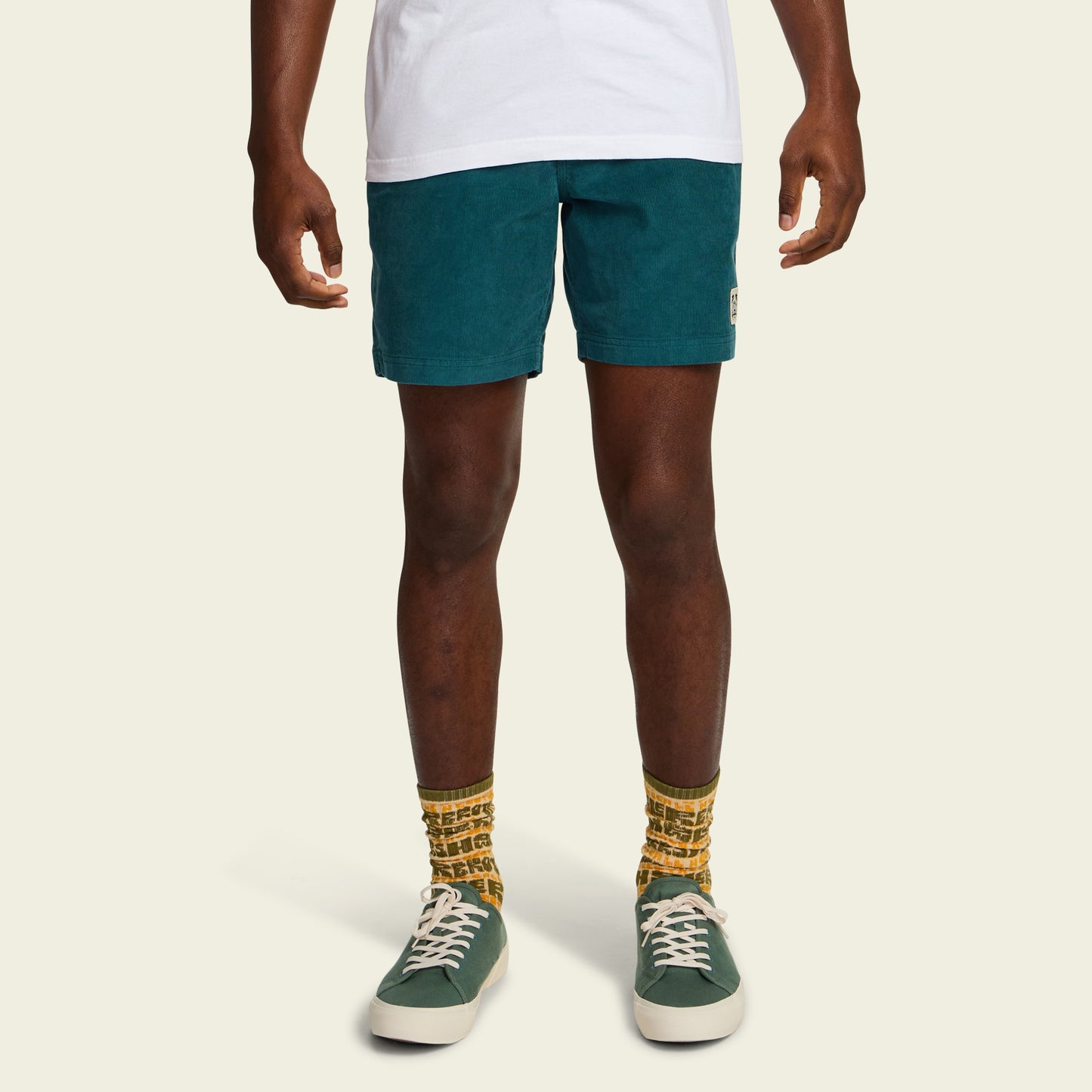 A person stands against a light background, wearing a white T-shirt, Pressure Drop Cord Shorts in teal—crafted from stretch cotton corduroy—yellow patterned socks, and green sneakers with white soles. Only the lower half of the body is visible.