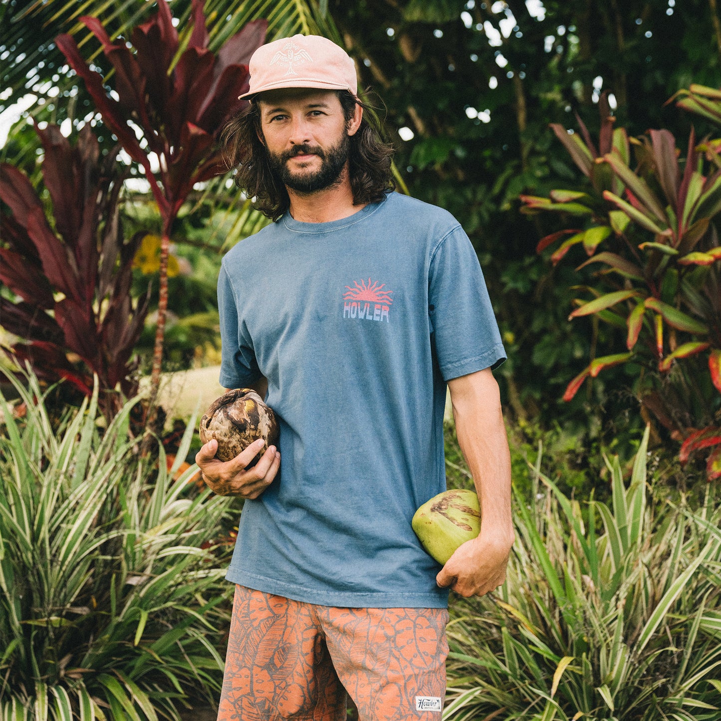 A man with long hair and a beard stands outdoors among tropical plants, wearing a vintage look blue cotton T-shirt, patterned shorts, and a pink cap. He holds two coconuts as he looks at the camera.