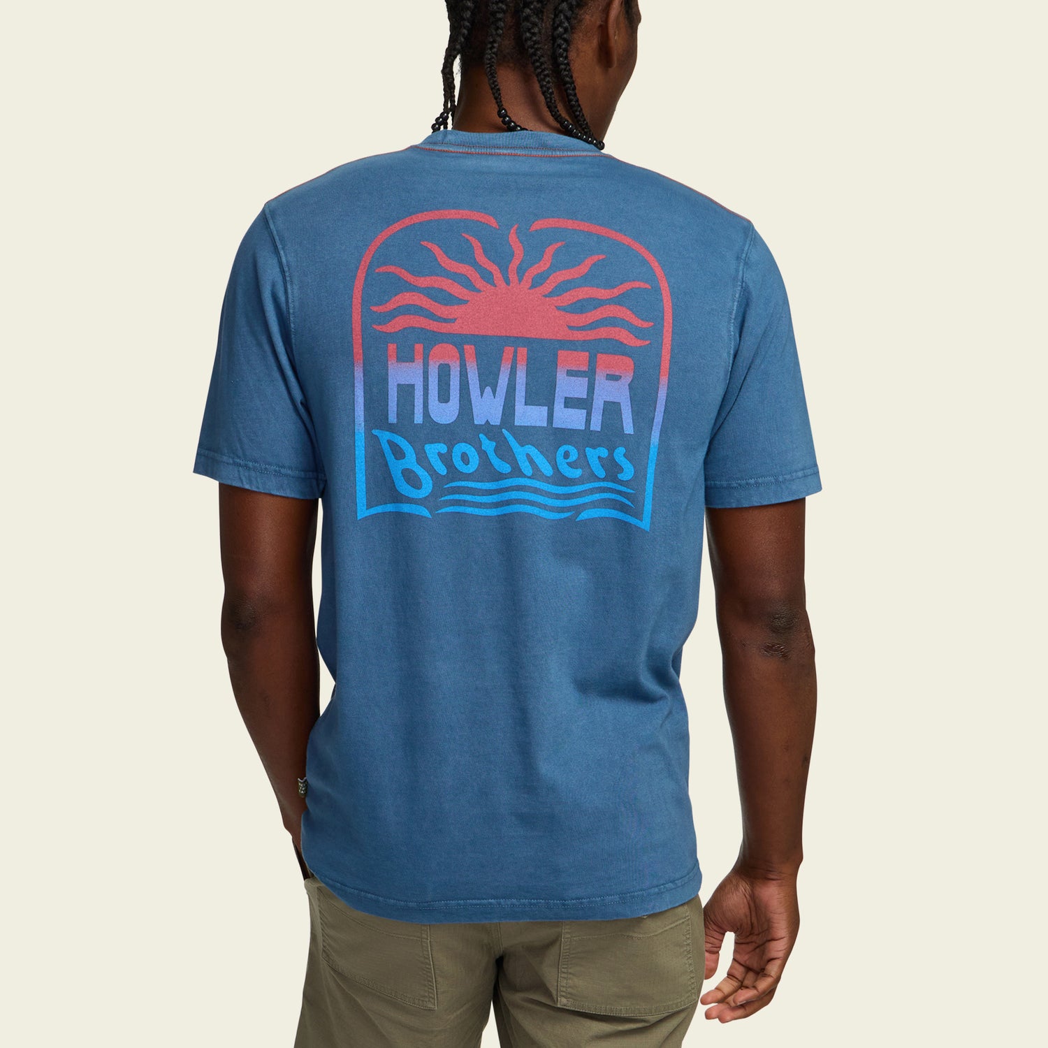A person wearing a resin-washed blue cotton t-shirt with a red sun design and HOWLER Brothers printed on the back in red, white, and blue, paired with khaki pants. The vintage look is completed as the person is seen from behind.