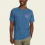 A man wearing a resin-washed blue short-sleeve cotton t-shirt with a small red and blue HOWLER graphic on the chest, and light green shorts, stands smiling against a plain light background for a vintage look.