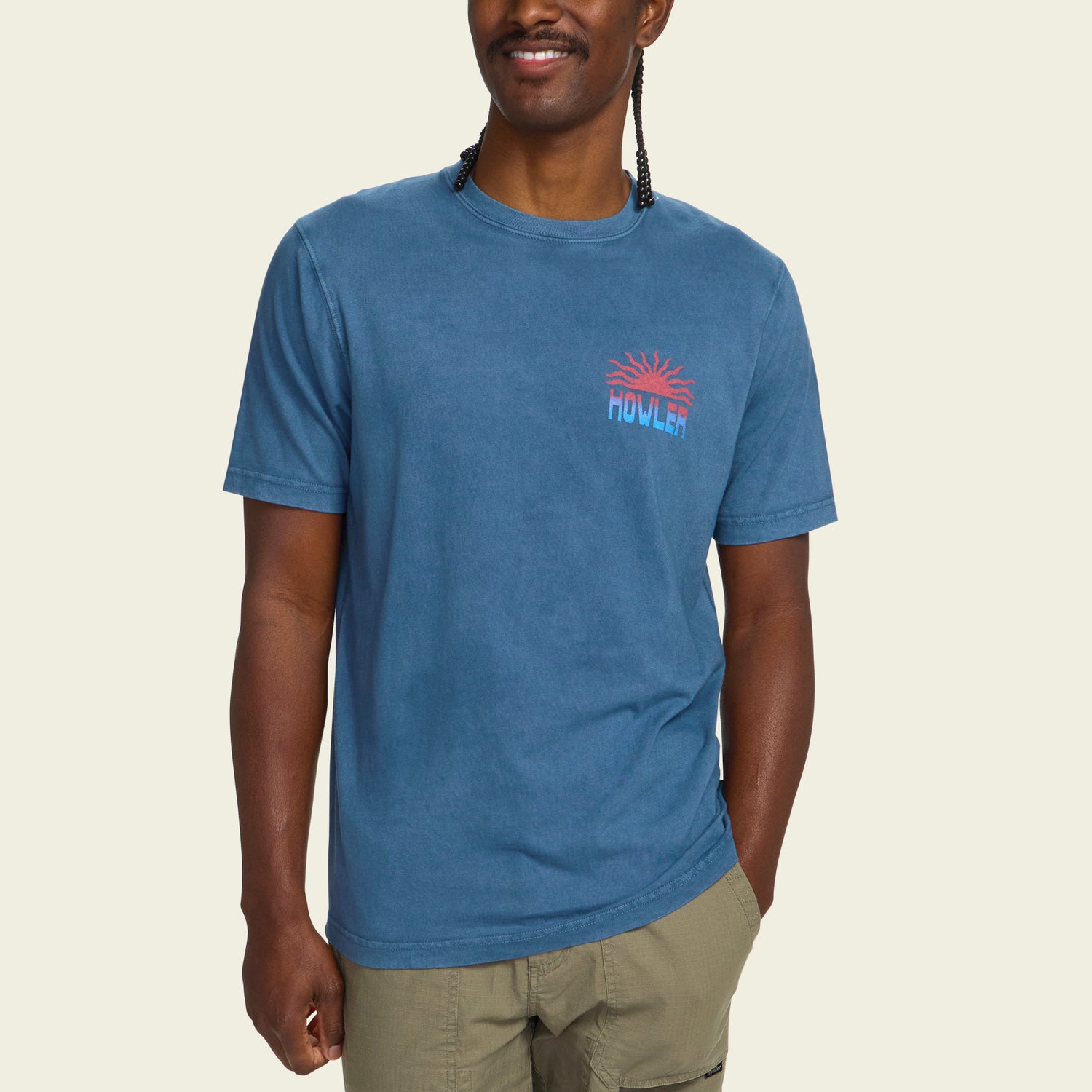 A man wearing a resin-washed blue short-sleeve cotton t-shirt with a small red and blue HOWLER graphic on the chest, and light green shorts, stands smiling against a plain light background for a vintage look.