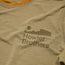 A close-up of a tan polyester spandex blend shirt with the Howler Brothers logo printed in dark brown, featuring stylized text below a minimalist landscape graphic with a sun and mountains.