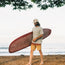 A man with long hair and a beard, wearing a beige cap, tan shorts, and a light green Howler blended t-shirt for maximum comfort, walks barefoot on a sandy beach carrying a red surfboard. The ocean and tree branches are in the background.