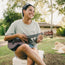 A smiling woman sits outdoors on a cooler playing a ukulele. She wears a Howler blended t-shirt and shorts, enjoying maximum comfort. Sunglasses rest on her head, with trees, a building, and a dog lying on the grass nearby.