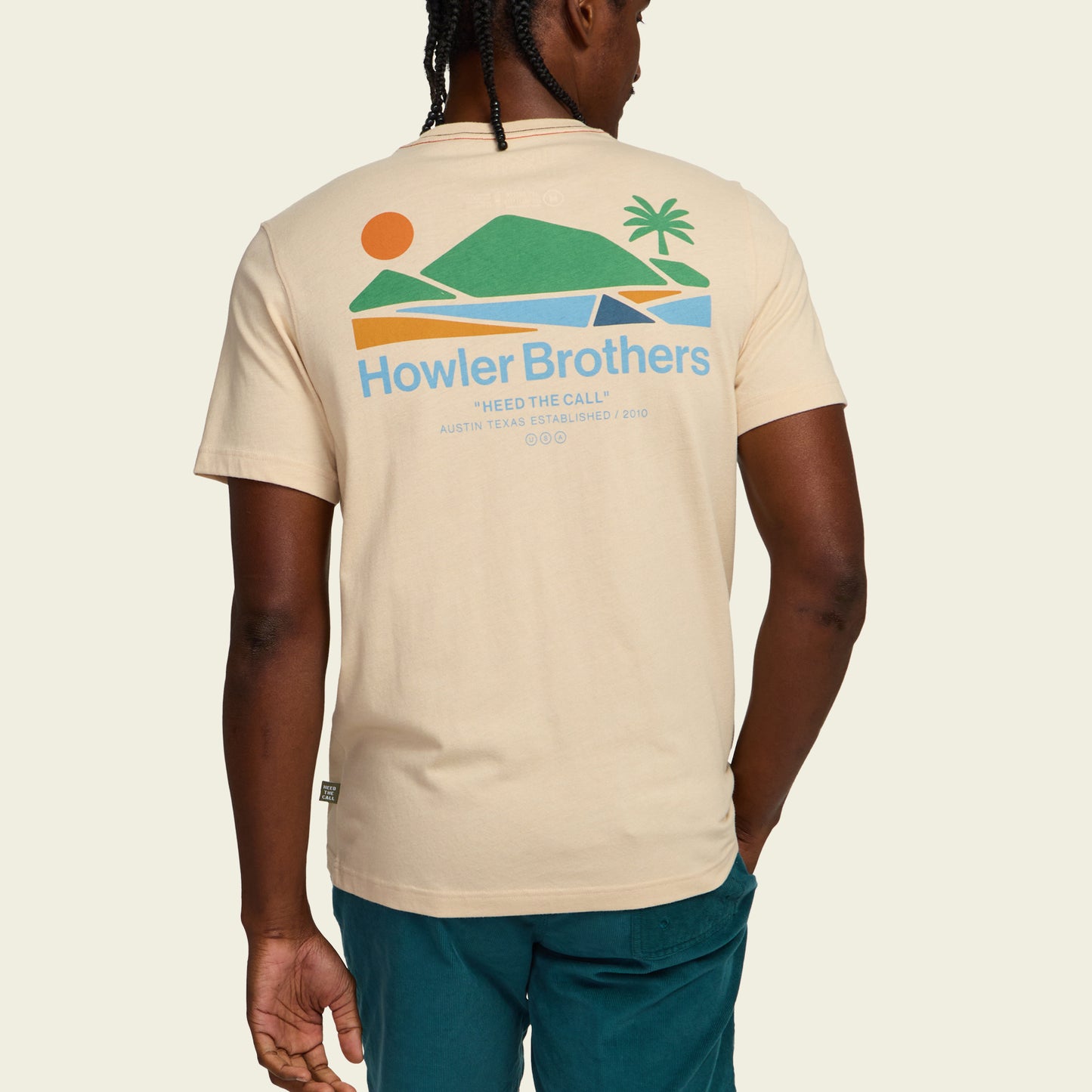 A man seen from behind wears a beige Howler blended t-shirt featuring a colorful graphic of a sunset, mountains, water, and palm tree, with the text Howler Brothers and Heed the Call, crafted for maximum comfort.
