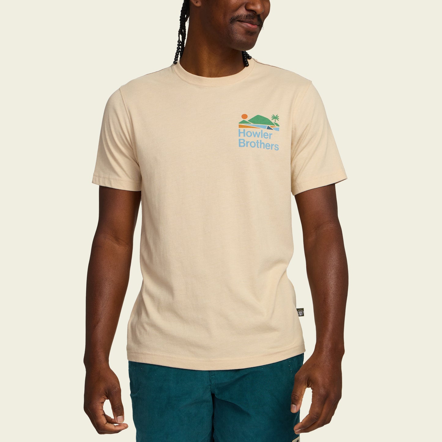 A person wearing a beige Howler blended t-shirt made from a soft cotton-poly blend for maximum comfort, featuring a small sun, wave, and palm tree graphic on the chest, paired with teal shorts against a plain background.
