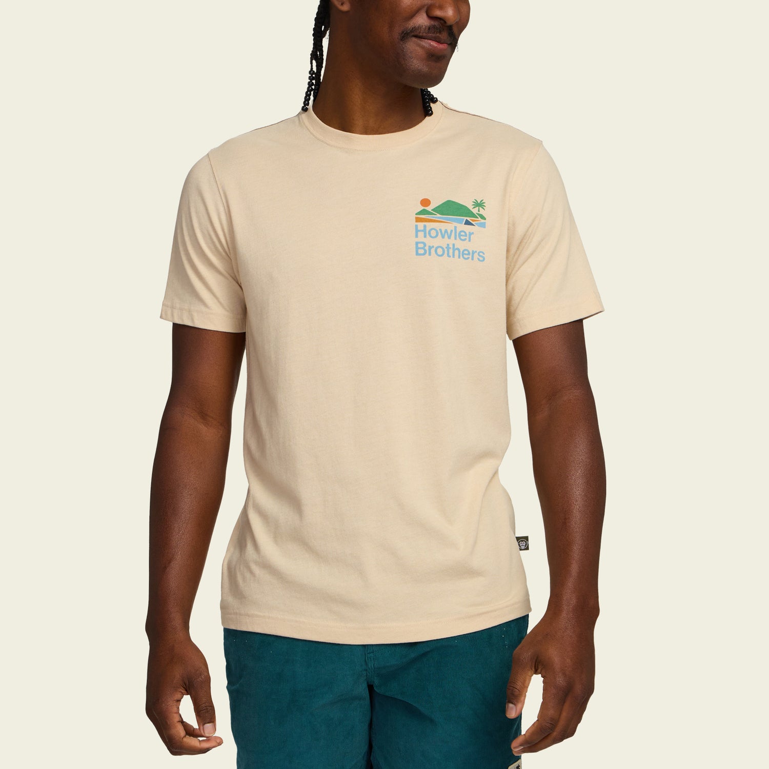 A person wearing a beige Howler blended t-shirt made from a soft cotton-poly blend for maximum comfort, featuring a small sun, wave, and palm tree graphic on the chest, paired with teal shorts against a plain background.