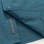 A close-up of a teal lightweight tech shirt with a snap button and a partially open flap, revealing a tag that reads “HEED THE CALL.”.