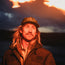 A man with long blond hair and a mustache wears a tech hat and dark jacket, standing outdoors at sunset with dramatic, glowing clouds in the sky behind him—capturing the Swan Valley Monster vibe.