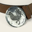 A round, silver El Gallo buckle from Cityboy Forge, embossed with a rooster and the words “HEED THE CALL,” attached to a brown leather belt.