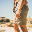 A person wearing versatile Horizon Hybrid Shorts made from ripstop fabric and a colorful patterned shirt stands outdoors on a sunny day, with rocks and a blue sky in the background. The image is focused on their lower torso and legs.
