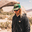 A person with long blonde hair wearing sunglasses, a green mesh cap, and a high pile fleece parka stands outdoors in a rocky, desert-like landscape under a partly cloudy sky.