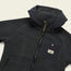 A dark gray outdoor jacket made from high pile fleece, featuring a hood, front zipper, zippered side pocket, and a contrasting black chest pocket with a Howler Brothers patch. The fleece parka is laid flat on a light background.