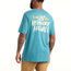 A man wearing a blue Howler Select T-Shirt with classic Howler designs and “Los Hermanos Howler” text on the back. Made from a soft cotton-poly blend, it features a fish graphic above the text. He stands with his back to the camera, tattoos visible on his right arm.