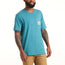 A man wearing a teal Howler Select T-Shirt, crafted from a soft cotton-poly blend, and tan pants stands against a plain white background. The shirt features classic Howler designs on the pocket, and the man has tattoos on both arms.