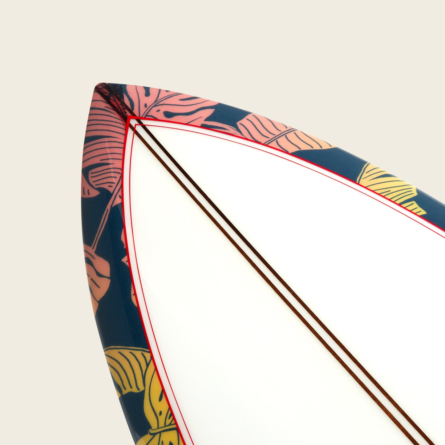 Close-up of the tip of an Almond Surfboards surfboard with a white center, red outline, and a dark blue border featuring a colorful tropical leaf pattern in pink and yellow. The background is a plain off-white.