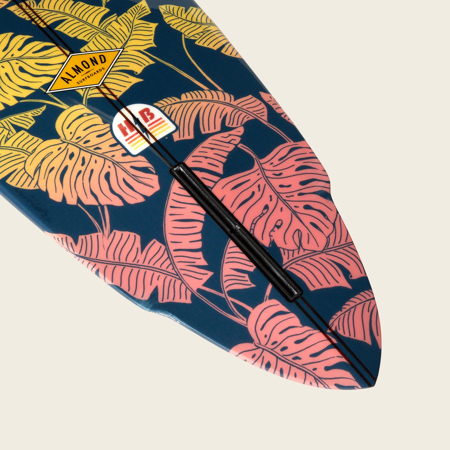 The tip of a surfboard decorated with pink and yellow tropical leaf patterns on a dark blue background, featuring brand stickers including one that says ALMOND from Almond Surfboards.