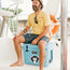 A man wearing a yellow t-shirt, patterned shorts, and sunglasses sits smiling on a blue YETI cooler adorned with heavy duty stickers and Howler Brothers stickers on a boat. A life preserver is visible in the background.