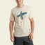 A man wearing a light beige Howler Select T-Shirt—crafted from a soft cotton-poly blend—with a graphic of a person holding a surfboard and the text BORN & AUSTIN RAISED, paired with dark shorts, stands against a plain light background.
