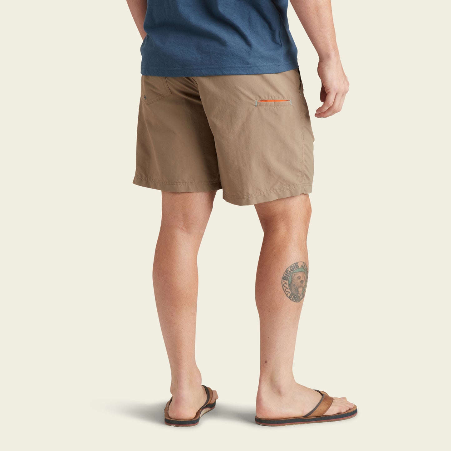 A person wearing a blue shirt, Horizon Hybrid Shorts made from ripstop fabric, and brown sandals is shown from the back. They have a round tattoo on their left calf. The background is plain and light-colored.