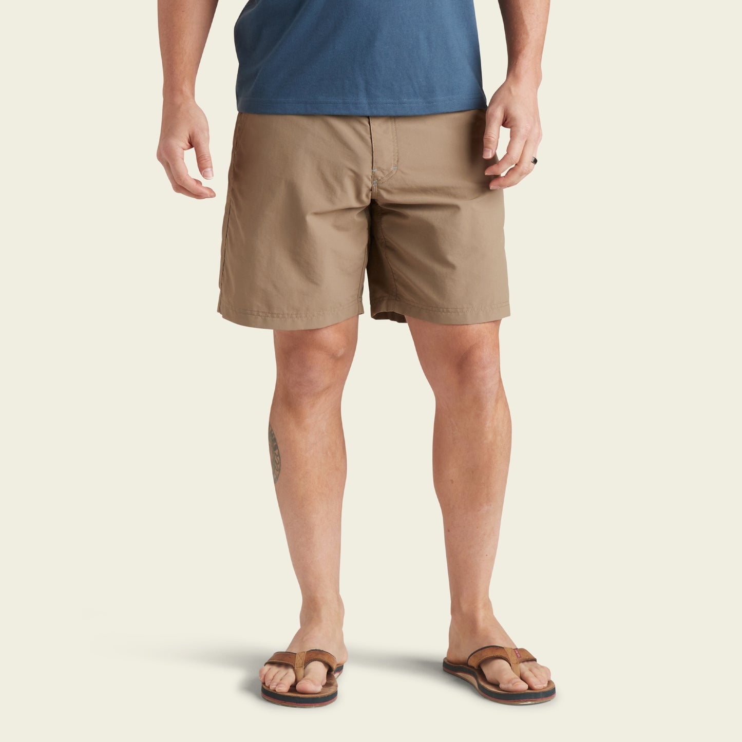 A person wearing a blue t-shirt and versatile shorts made from ripstop fabric with brown flip-flops stands facing forward. Only their body from the shoulders down is visible against a plain light background.
