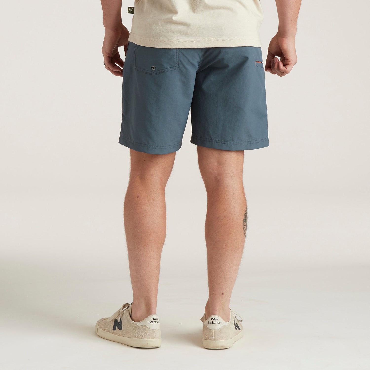 A person stands facing away, wearing a cream T-shirt, white sneakers, and versatile blue Horizon Hybrid Shorts with a back pocket made from durable ripstop fabric. The background is plain and light-colored.