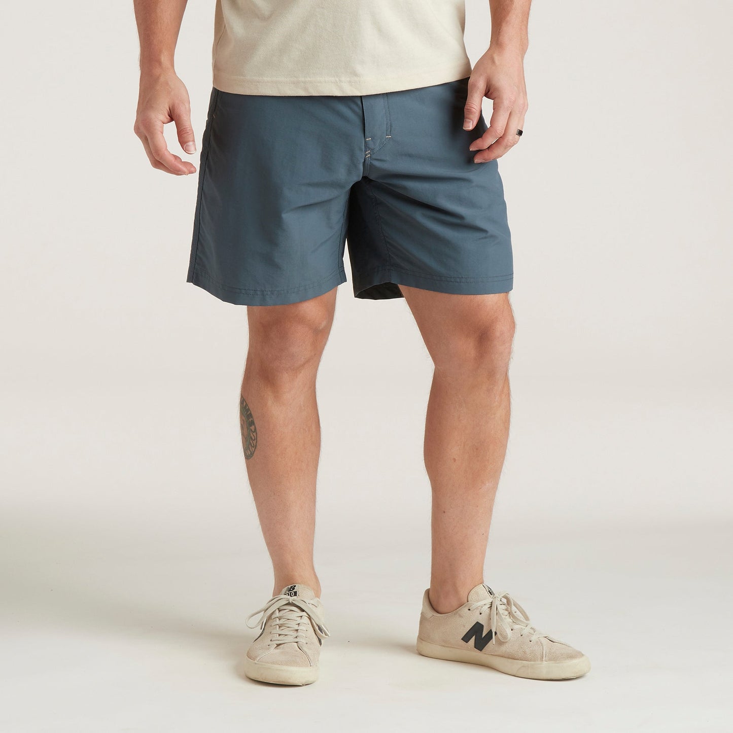 A person stands against a plain background wearing a beige T-shirt, Horizon Hybrid Shorts in blue ripstop fabric, and beige sneakers. Their hands are relaxed at their sides, and a round tattoo is visible on their left leg.