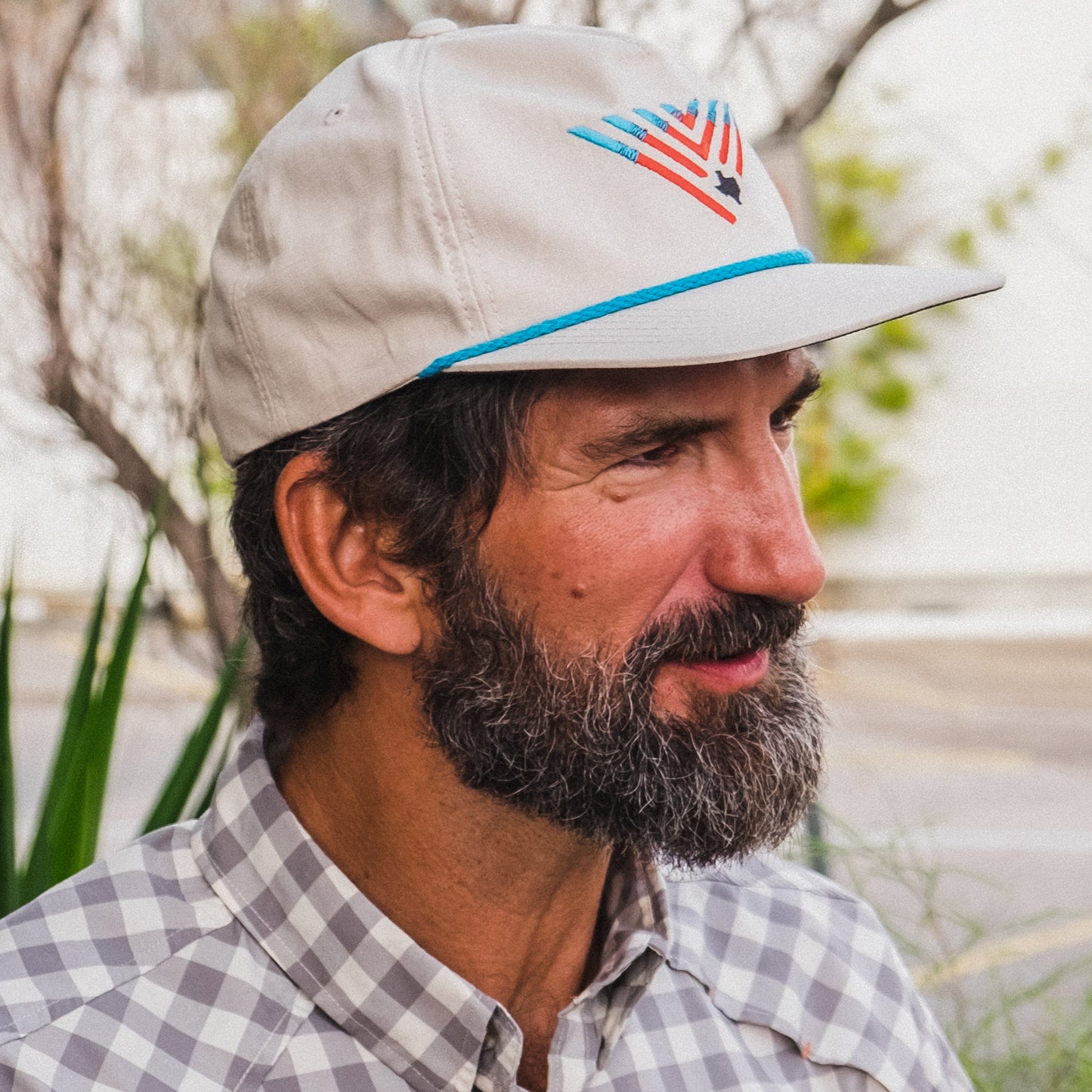 A man with a thick beard and mustache wears an unstructured snapback featuring a dark under brim and adjustable snap back closure, paired with a plaid shirt, standing outdoors amid greenery and a blurred background.