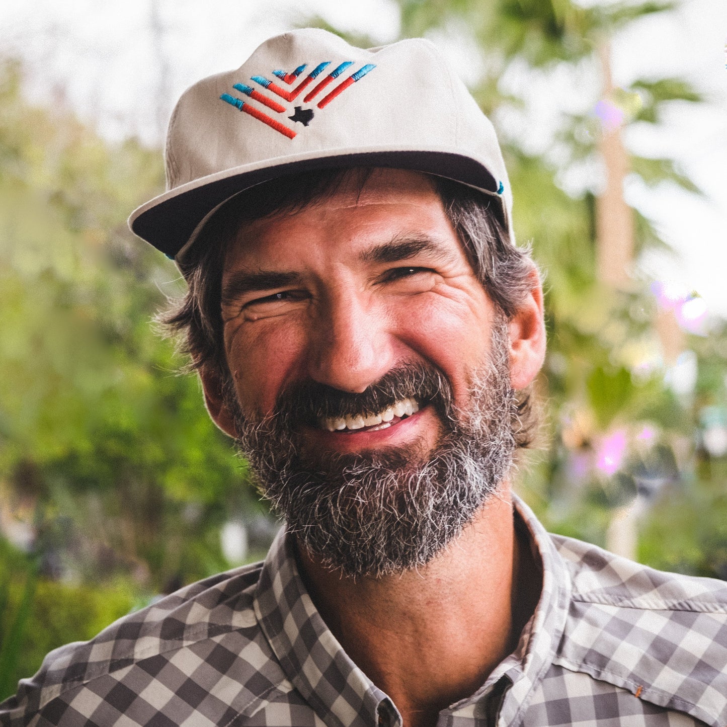 A bearded man wearing a patterned shirt and a light-colored fishing hat with a colorful design and adjustable snap back closure smiles outdoors, with green foliage in the blurred background.