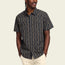 A man wears a short-sleeve, button-up H Bar B Snapshirt featuring a vertical floral pattern in beige and gold tones on a dark background, paired with light-colored pants, standing against a plain light background.