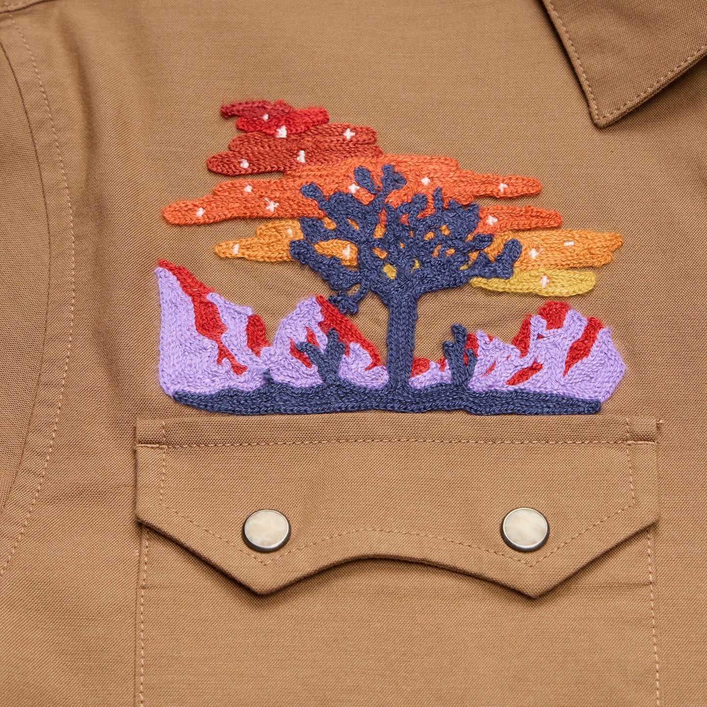 A brown Crosscut Deluxe jacket features vintage inspired embroidery of a tree with purple mountains, an orange, yellow, and red sunset, and small white stars above a buttoned chest pocket.