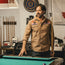A man with dark hair and a beard stands by a pool table, smiling. He wears a tan Crosscut Deluxe shirt with vintage inspired embroidery. Pool cues and various items are visible in the background on the wall and shelves.