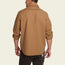 A man stands with his back to the camera, wearing a light brown Crosscut Deluxe long-sleeve shirt with rolled-up sleeves and black pants, featuring vintage inspired embroidery against a plain, light background.
